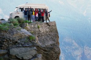 A group of friends celebrating on a cliff edge with a van in the background during a mountain adventure.